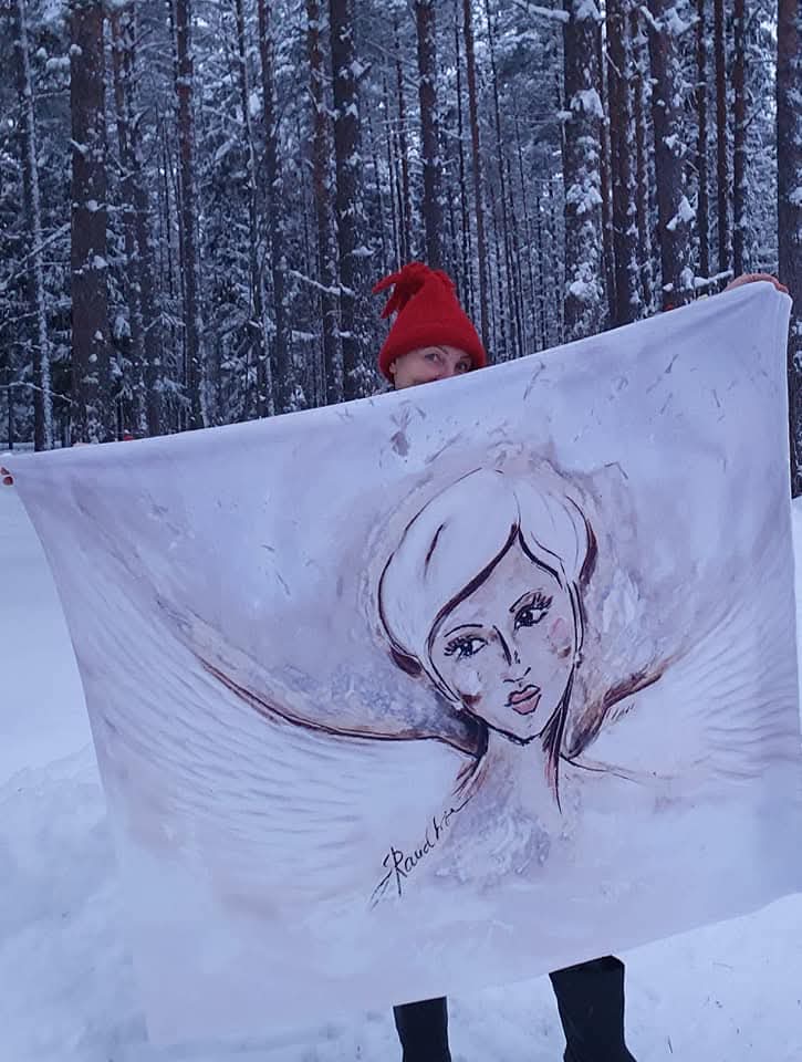 Punase mütsiga inimene hoiab lumises männimetsas suurt valget salli, millele on trükitud Inglitar kollektsiooni tiibadega naisenägu.