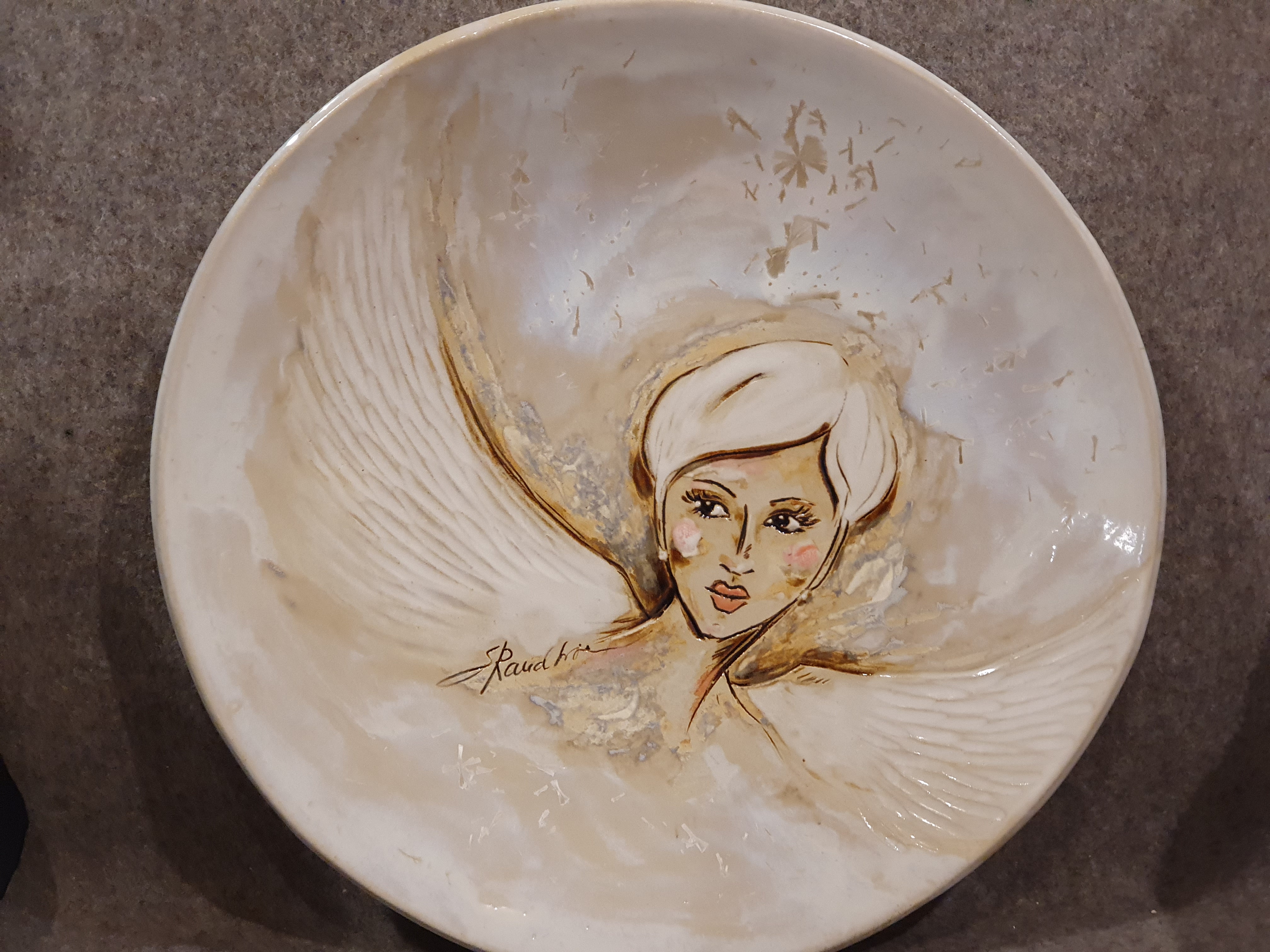 Ümmargune heledates toonides keraamiline vaagen. Keskel on maalitud ja voolitud lühikeste heledate juustega naise nägu ning suured, detailsed ja reljeefsed ingli tiivad. All on näha kunstnik Raudkivi signatuuri.